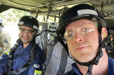 Two men wearing helmets, protective eyewear and blue jumpsuits are seated inside a military helicopter.